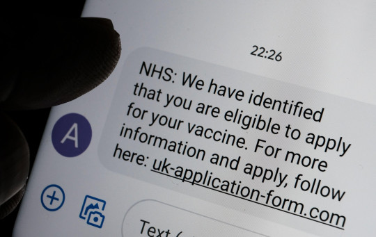 2E2DGMX Stafford, United Kingdom - January 13 2021: Scam coronavirus vaccine text message seen on the smartphone screen and blurred silhouette of finger point
