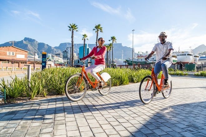 A vacationing man and woman riding bikes down a sunny street with palm trees and mountains behind the town.