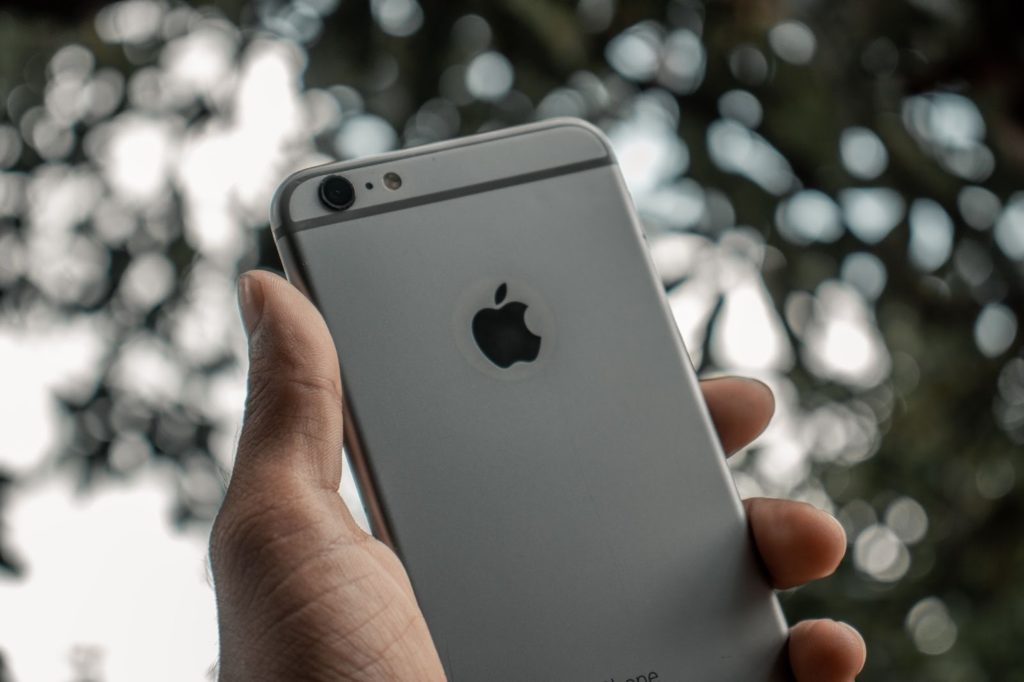 A picture of a hand holding up a grey iPhone with the camera side facing the viewer, against a blurred background. 
