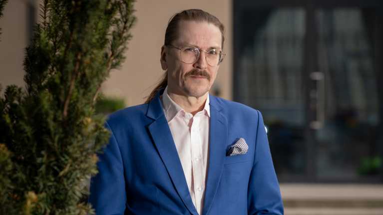 Portrait of a man with glasses, a mustache and dark hair combed back into a ponytail, wearing a blue blazer.
